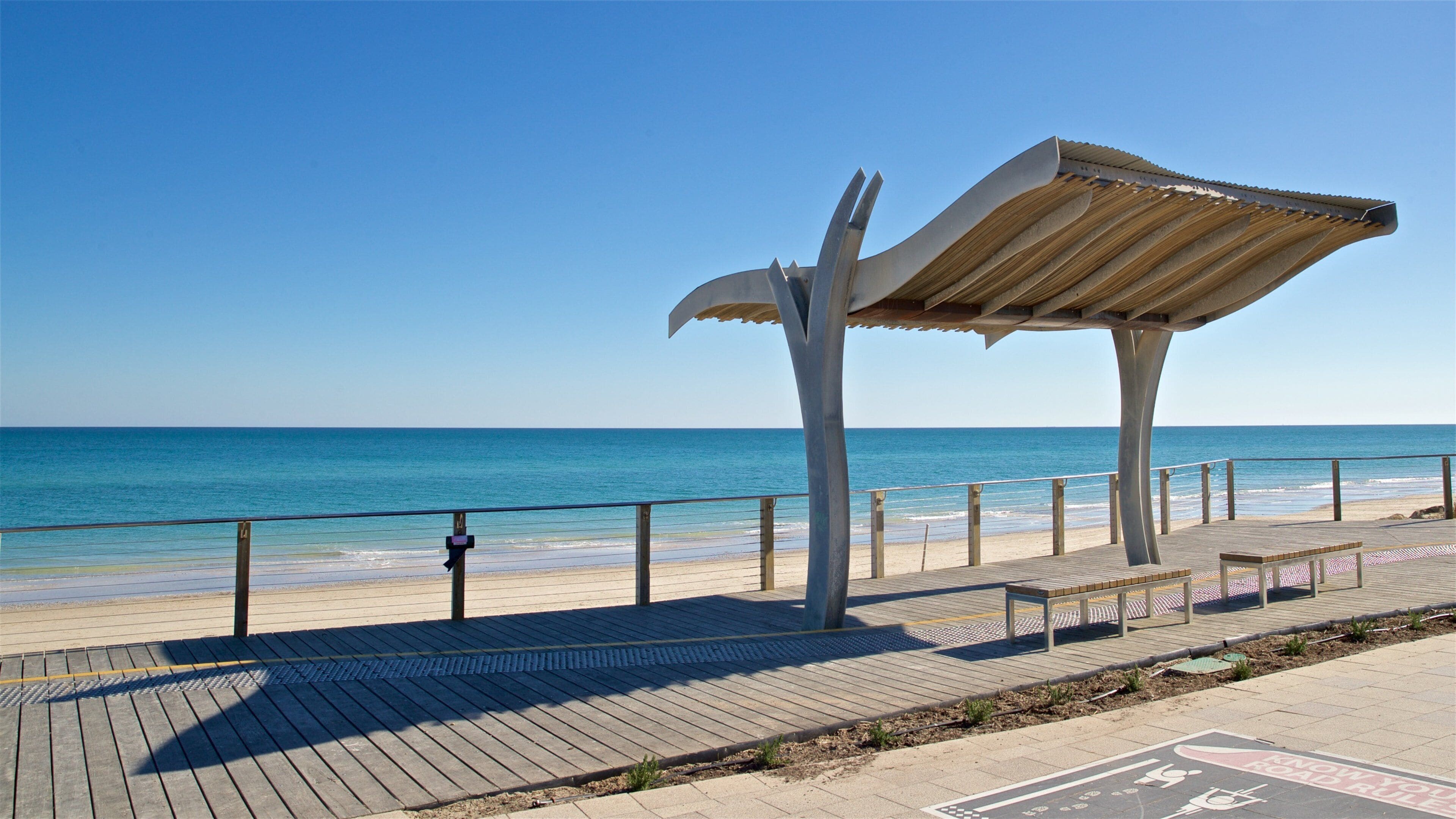 Una playa con un paseo marítimo de madera, barandilla de metal y una escultura de metal.
