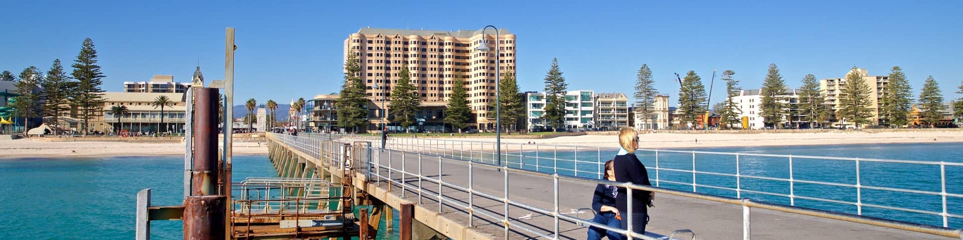 A pier extending into turquoise water, with a beach and buildings in the background.
