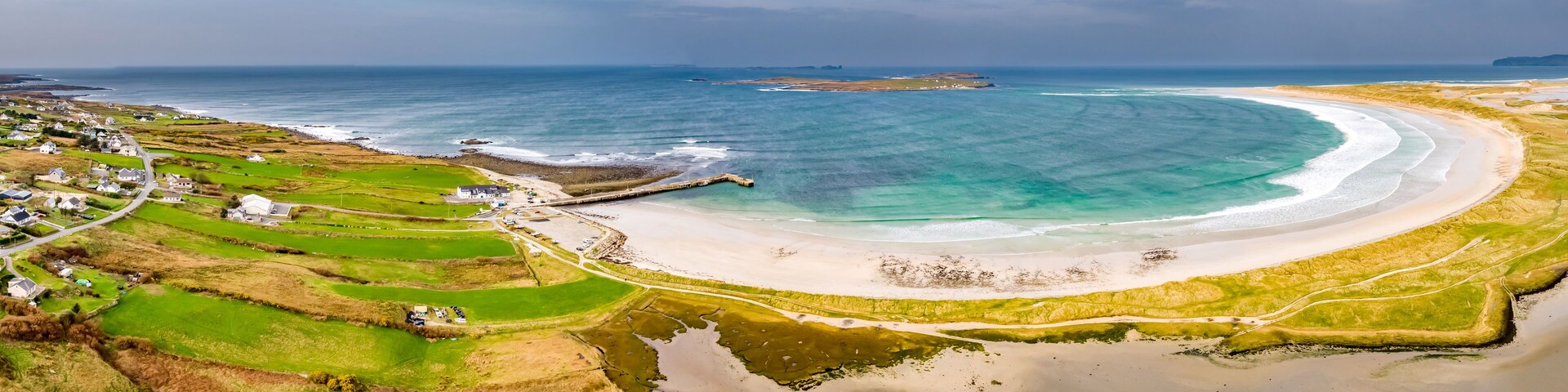 Paisagem costeira com uma praia de areia, campos verdes e um pequeno cais.