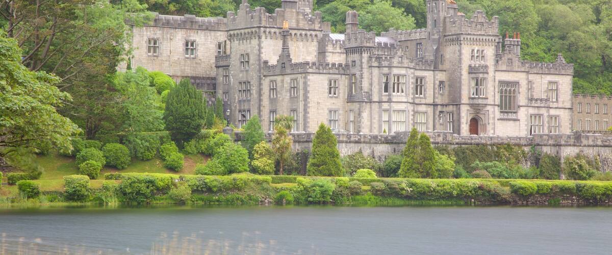 A large, historic stone castle surrounded by lush greenery and a calm lake.