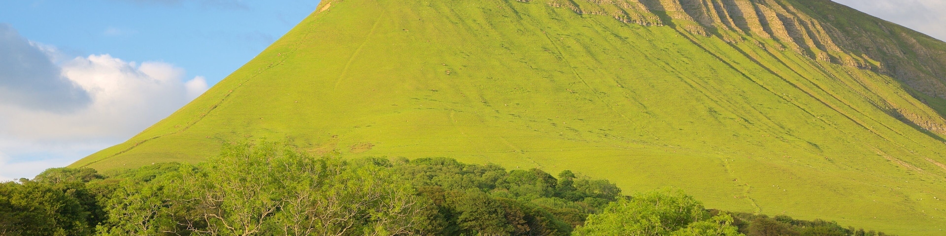 A large green hill with a steep cliff, surrounded by trees and a small house.