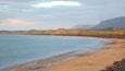 A coastal landscape with a sandy beach, calm sea, dunes, and distant mountains.