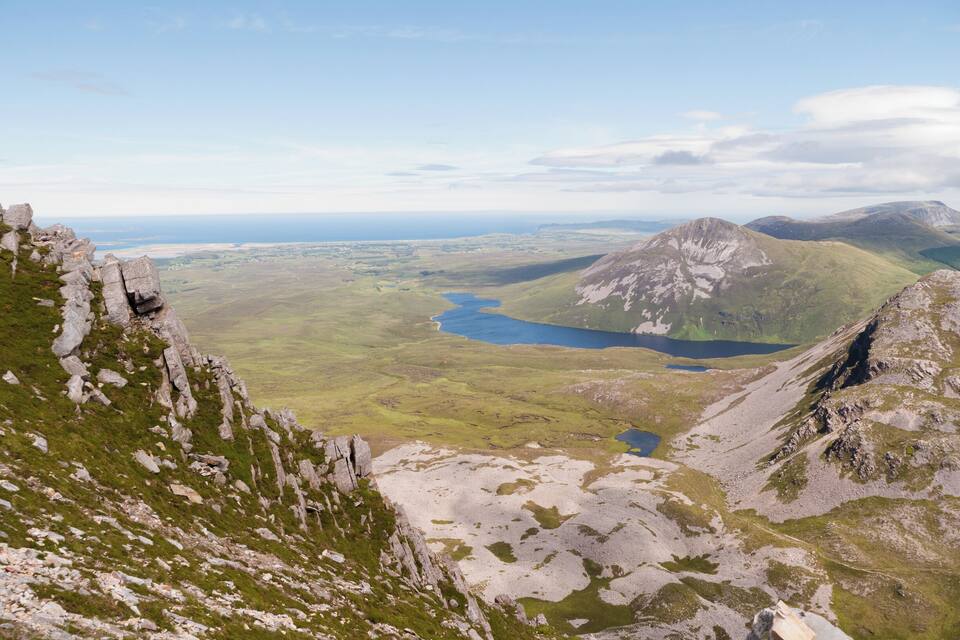 Un paisaje montañoso con afloramientos rocosos, un valle con un lago y colinas distantes.
