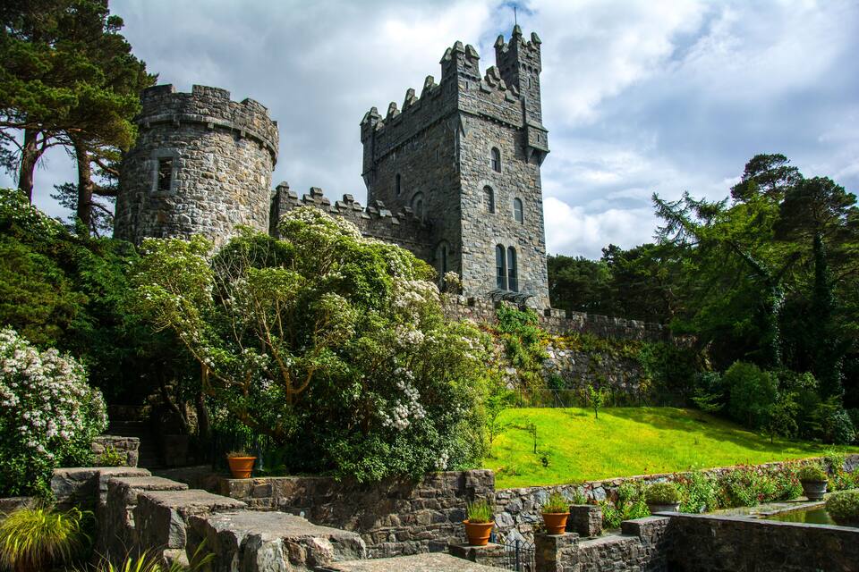Un castillo de piedra histórico con una torre, rodeado de abundante vegetación y plantas en flor.