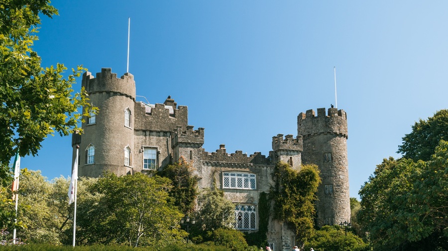 Un château historique, doté de deux tours imposantes, entouré d’une végétation luxuriante et surplombant un ciel d’un bleu éclatant.