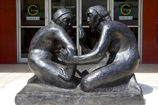 Eine Skulptur von zwei Figuren in nachdenklicher Pose vor einem Gebäude mit der Beschilderung 'National Gallery of Jamaica'.