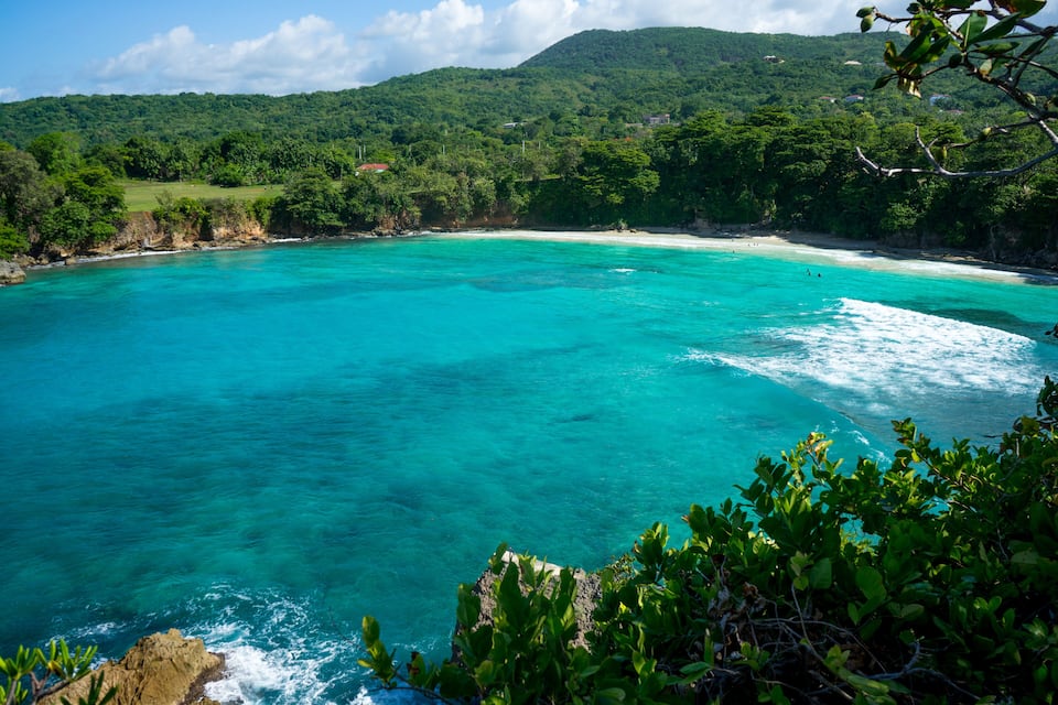 A turquoise coastal bay with clear waters, surrounded by lush green hills and a rocky coastline.