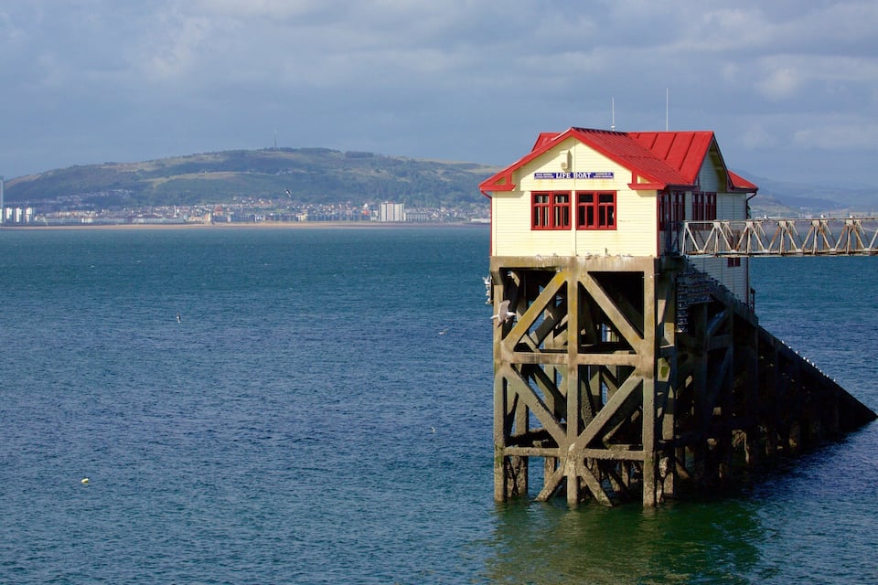Um farol sobre um cais com telhado vermelho, com vista para um mar calmo e um horizonte urbano distante.