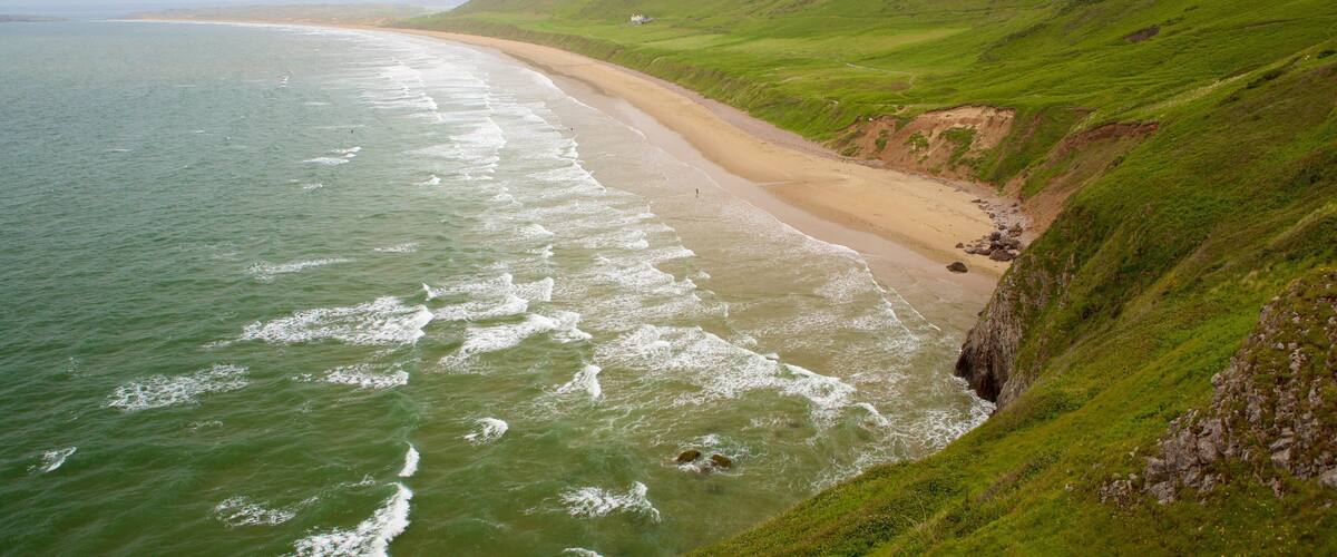A coastal landscape with a sandy beach, green hills, and the ocean.