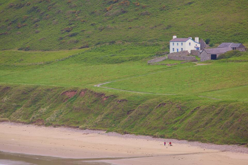 Paisaje costero con una casa blanca sobre una colina cubierta de hierba, una playa de arena y algunas personas caminando.