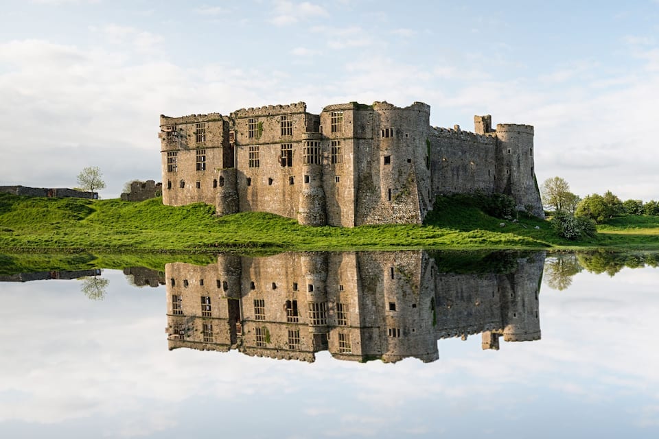 Eine historische Burg mit einem Burggraben, umgeben von üppiger Vegetation und spiegelnd im ruhigen Wasser.