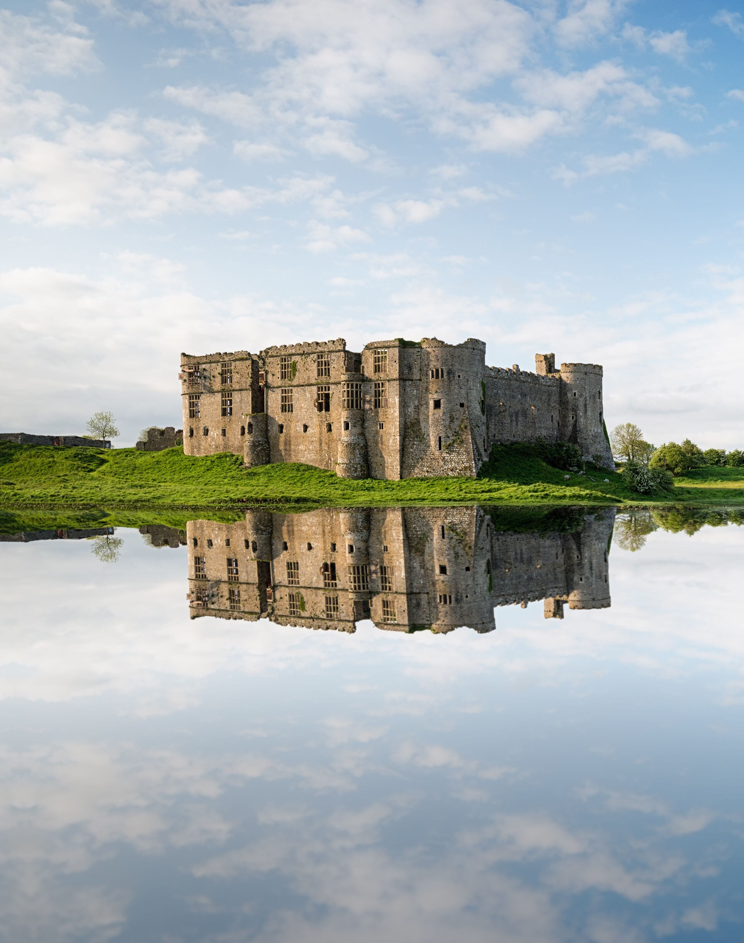 A historic castle with a moat, surrounded by lush greenery and reflecting in calm water.