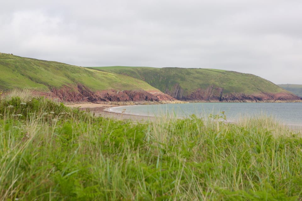 A coastal landscape with grassy hills, a sandy beach, and calm water.