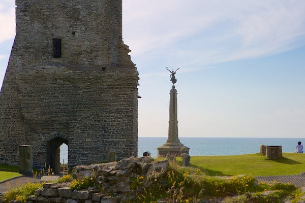 Une tour de pierre, un monument surmonté d’une statue, et une personne assise dans l’herbe, près de la mer.