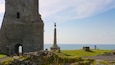 A stone tower, a monument with a statue, and a person sitting on the grass by the sea.