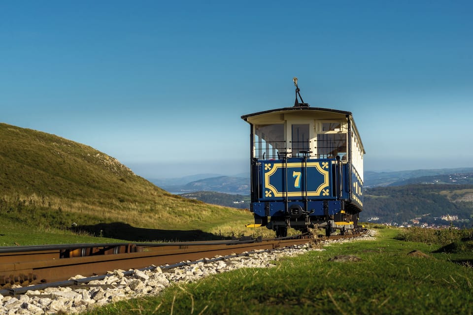 Um vagão de trem azul e amarelo com o número 7 em uma linha férrea, cercado por colinas verdes e um céu azul límpido.
