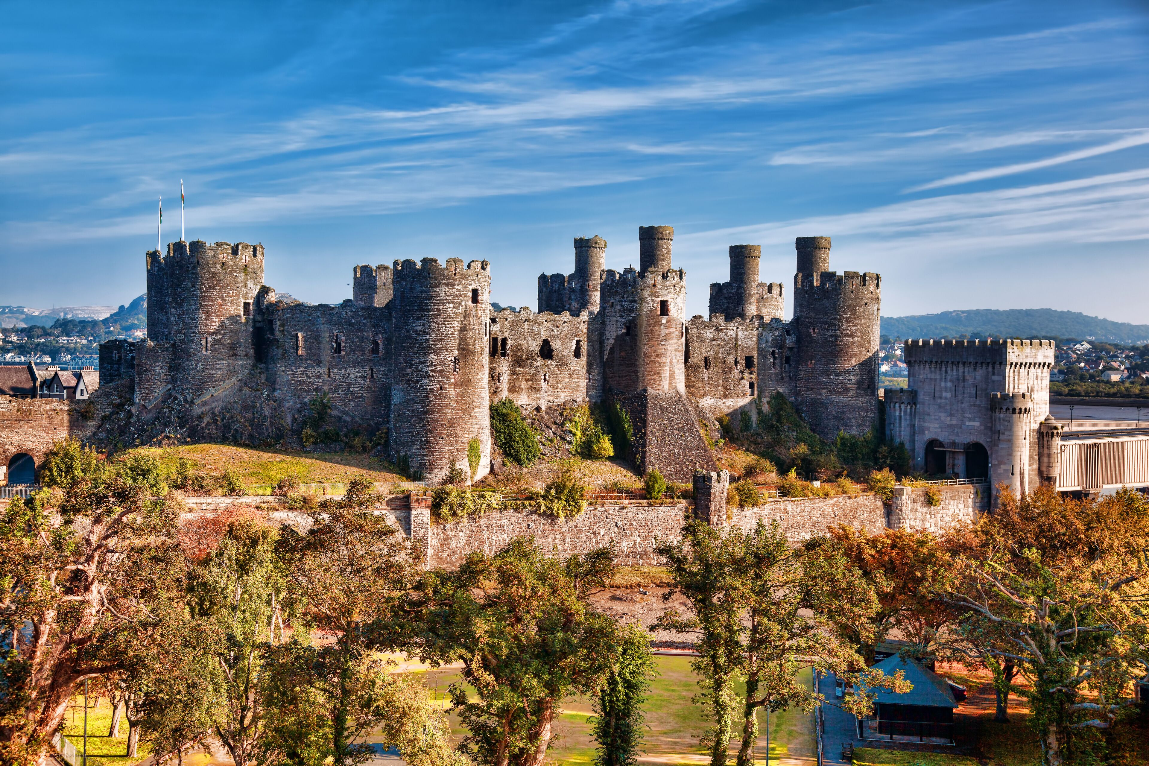 A historic castle with multiple towers and a surrounding wall, set against a backdrop of hills and trees.