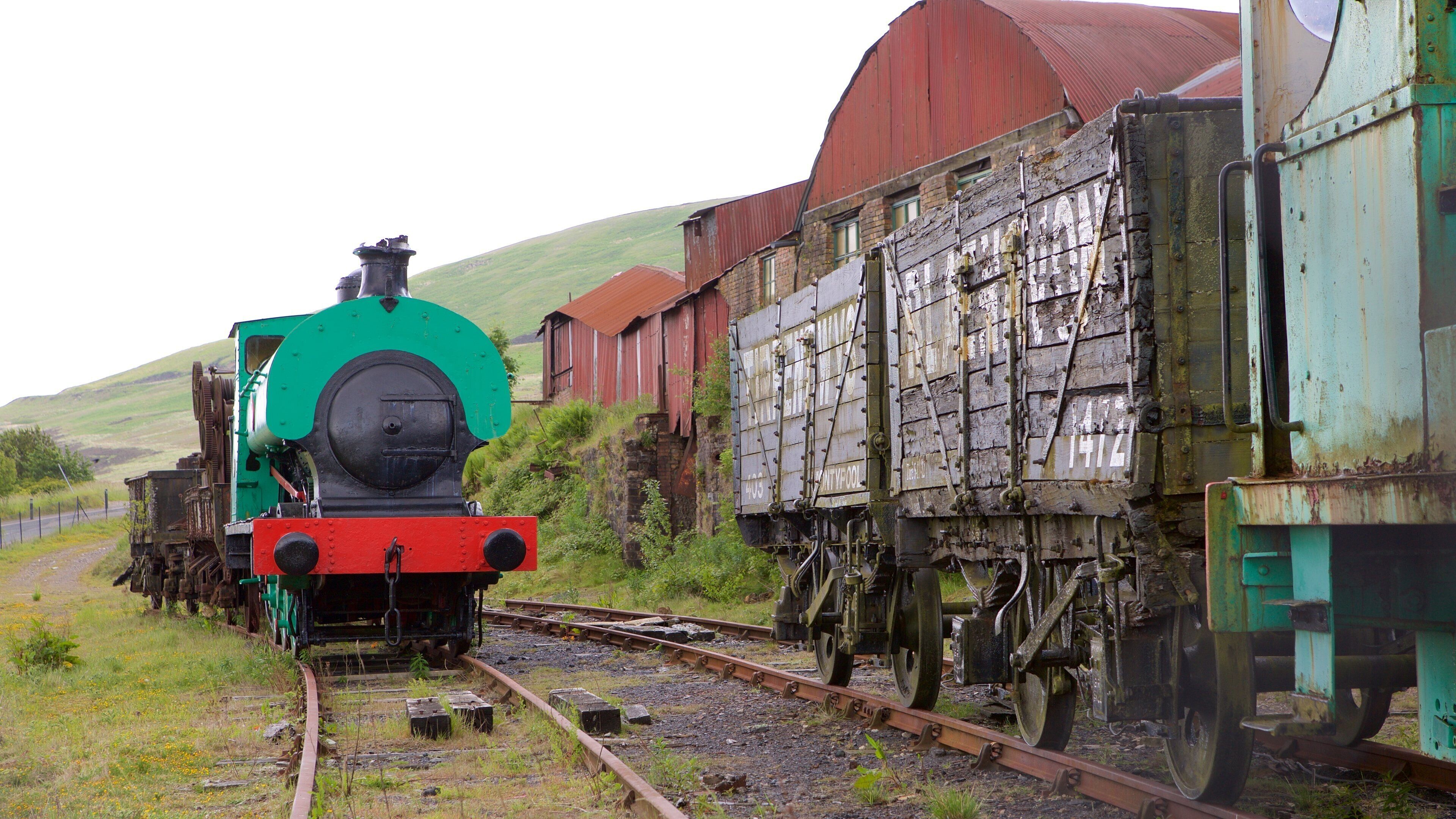 Una locomotora de vapor verde y roja arrastra un tren de carga con un vagón de freno de madera, pasando junto a un granero rojo y una colina al fondo.