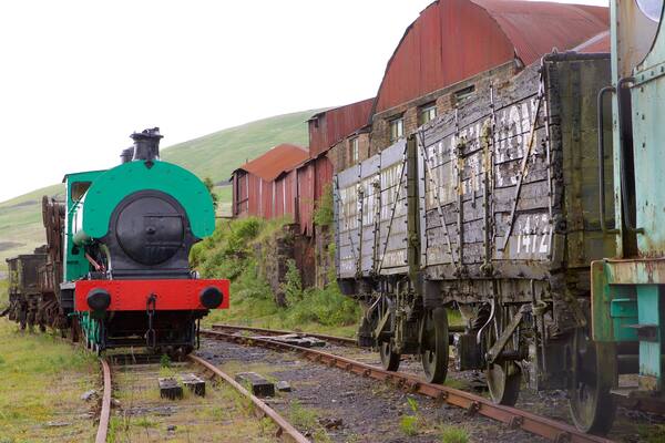 A green and red steam locomotive pulling a freight train with a wooden caboose, passing by a red barn and a hill in the background.