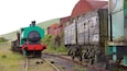 Eine grüne und rote Dampflokomotive zieht einen Güterzug mit einem hölzernen Waggon, vorbei an einer roten Scheune und einer Hügelkulisse im Hintergrund.