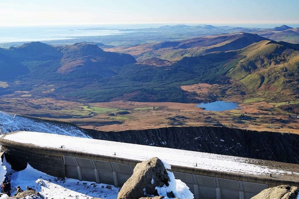 Uma cordilheira nevada com um lago distante e colinas ondulantes.