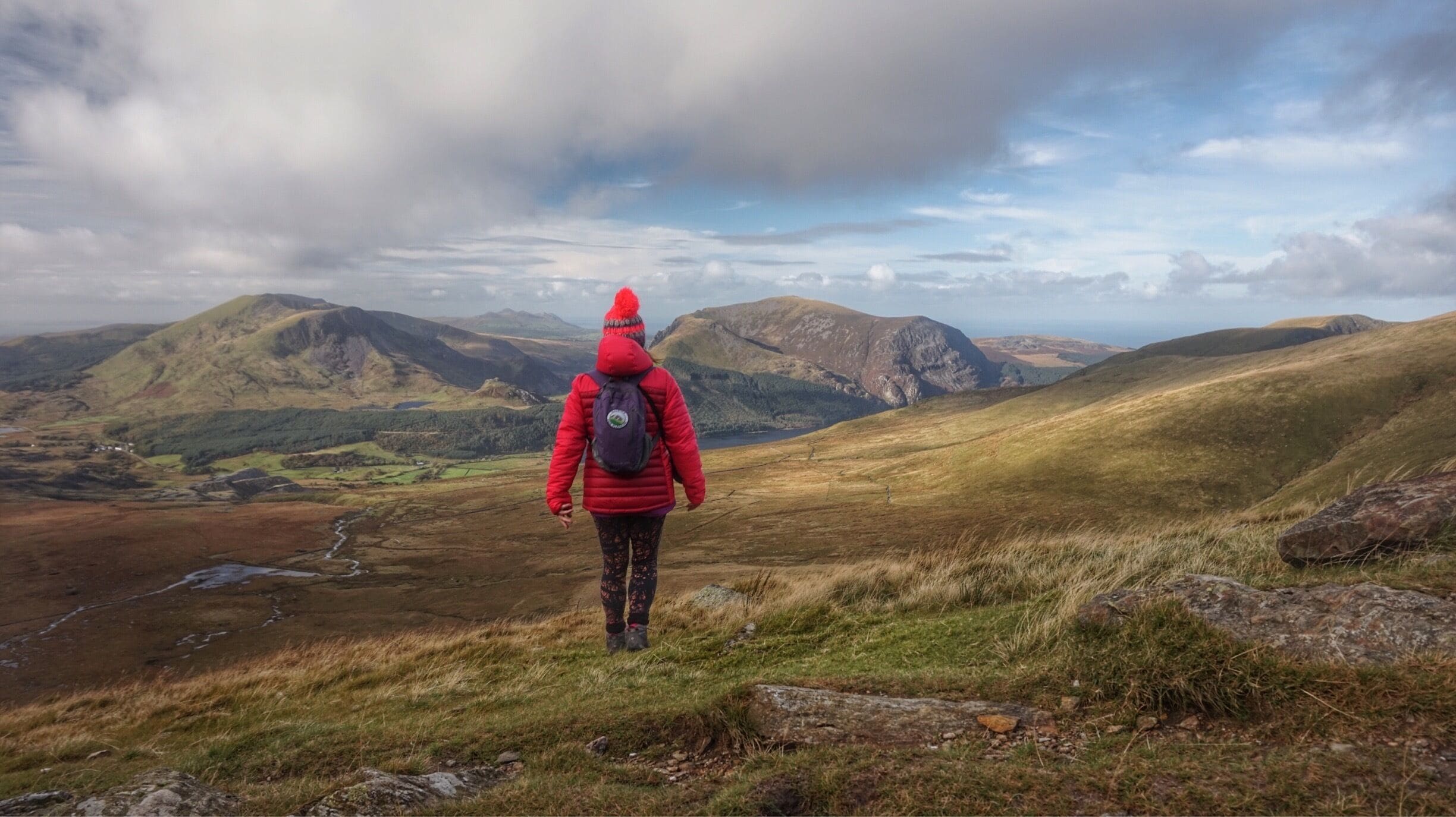 A hiker in a red jacket and hat walking on a grassy hillside with mountains in the background.