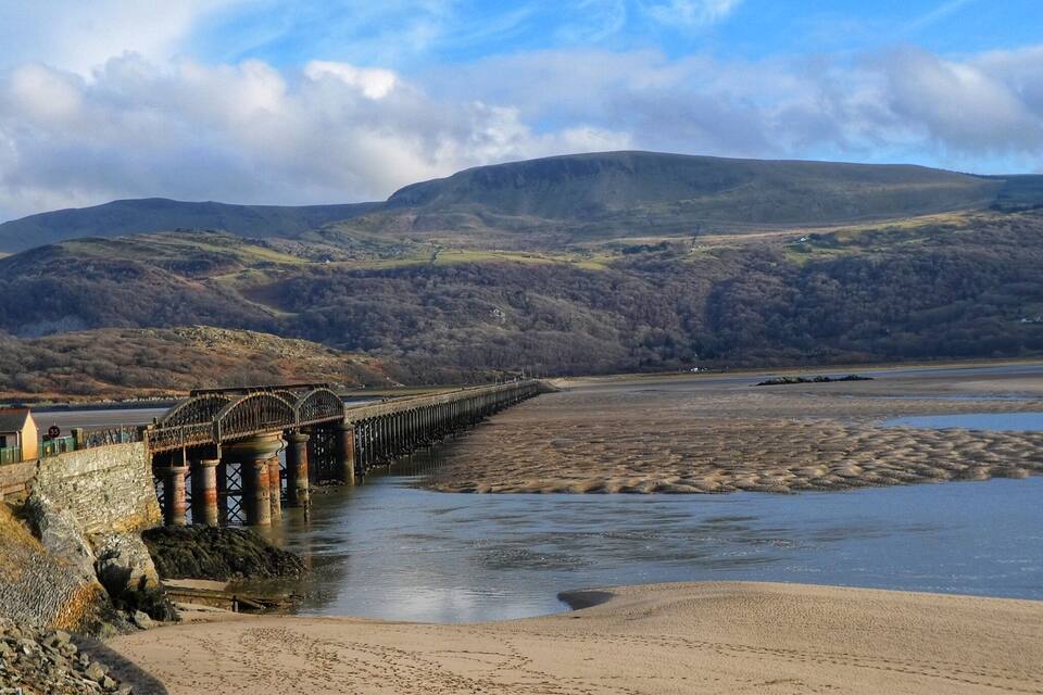 A coastal landscape with a bridge over a river, sandy beach, and hills in the background.