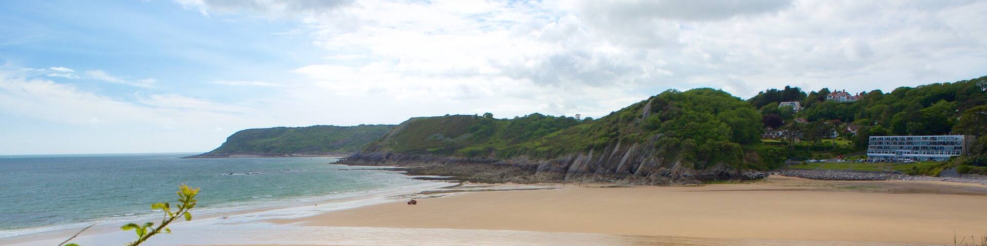 A wide sandy beach with gentle waves, green hills in the background, and a few buildings on the hillside.