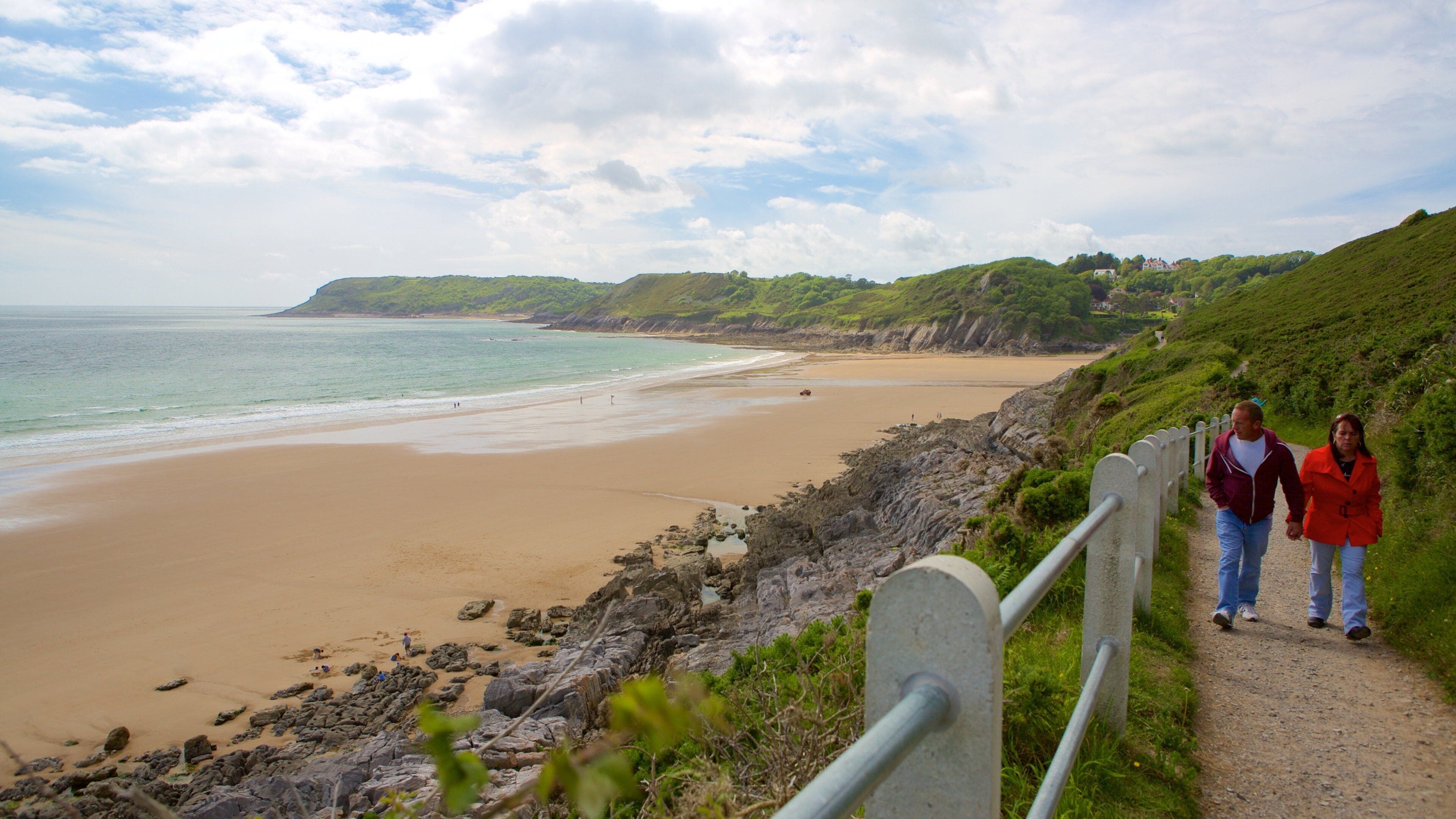 A coastal path with a sandy beach, rocky shoreline, and green hills.