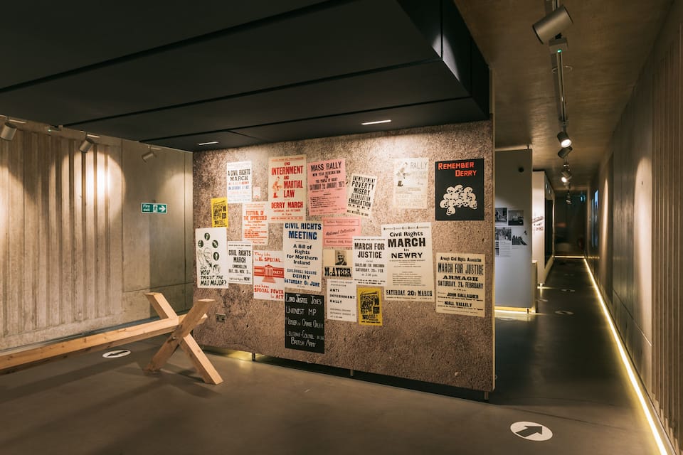 A corridor with a wall covered in historical posters and signs, a fire exit sign, and a wooden structure resembling a chair.