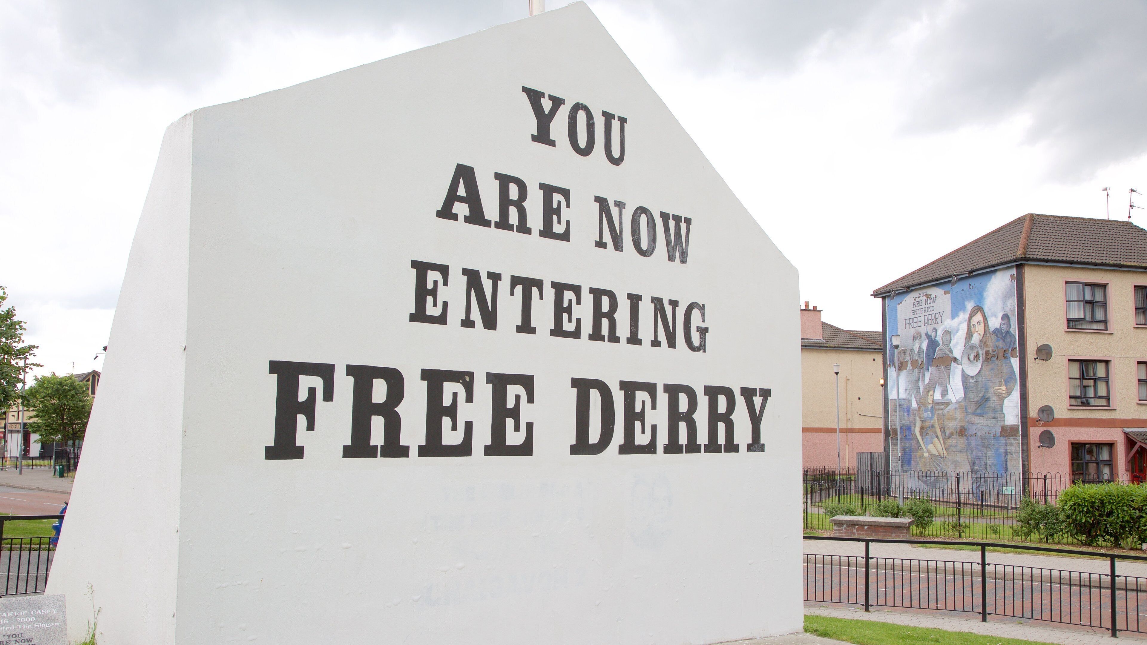 A large white sign with black lettering that reads "YOU ARE NOW ENTERING FREE DERRY." In the background, there is a building with a mural and a fence.