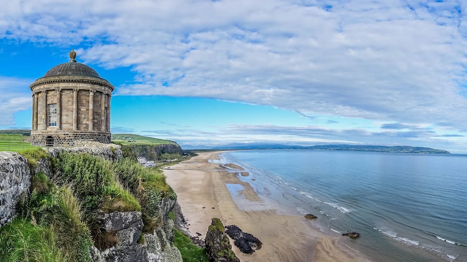 A coastal landscape with a historic stone tower, a sandy beach, and a view of the sea and distant hills.