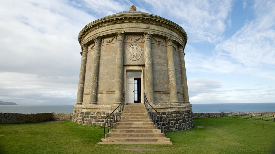A stone structure with a dome and columns, surrounded by grass and a stone wall, overlooking the sea.