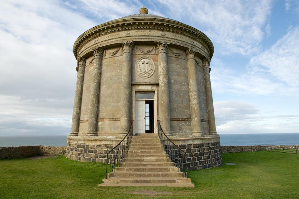 A stone structure with a dome and columns, surrounded by grass and a stone wall, overlooking the sea.
