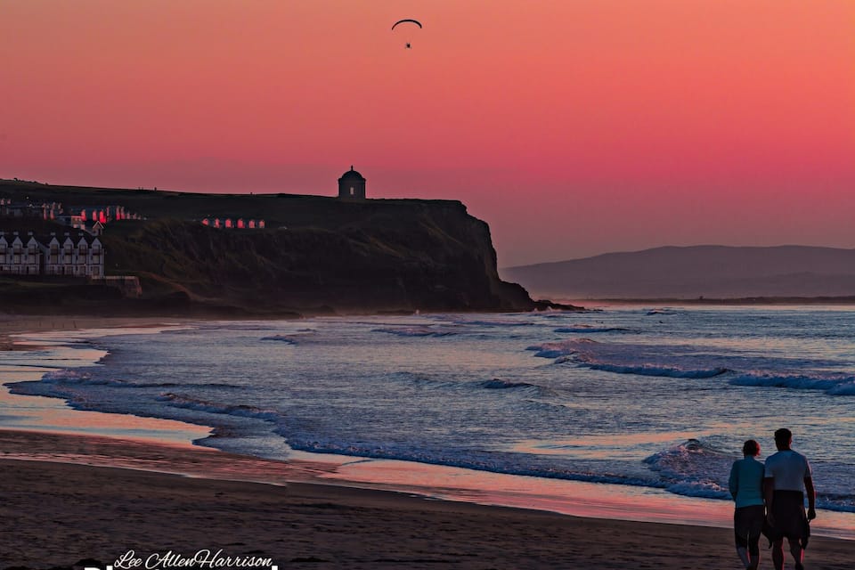 A beach with waves, a lighthouse, and a person walking.