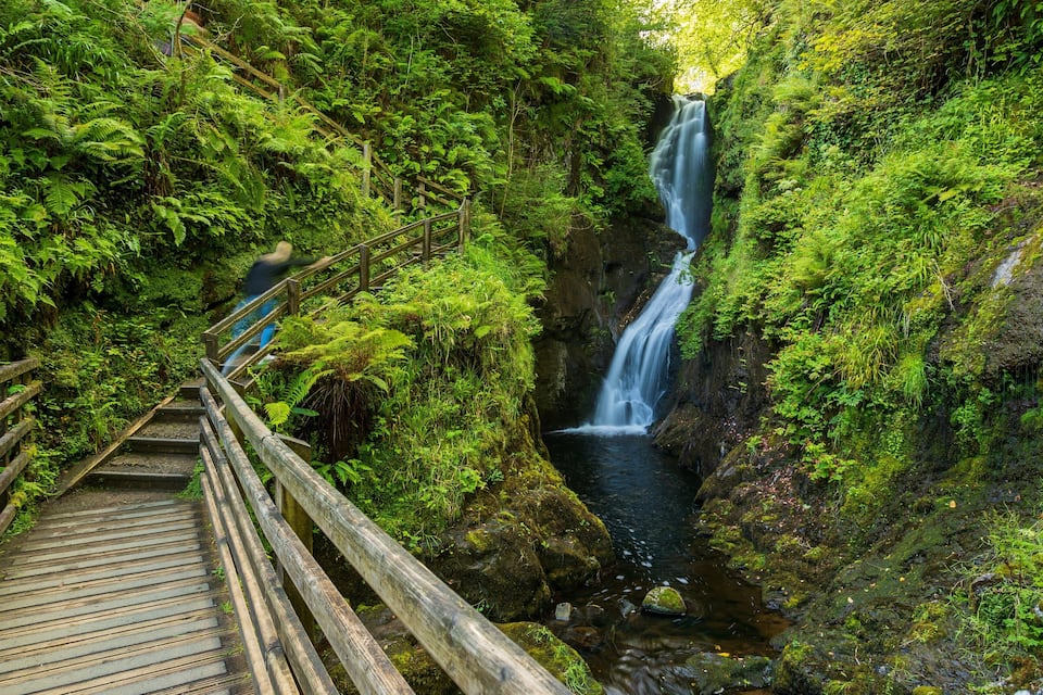 A wooden bridge leading to a waterfall surrounded by lush greenery.