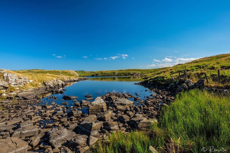 A calm lake surrounded by rocky terrain and grassy hills under a clear blue sky.