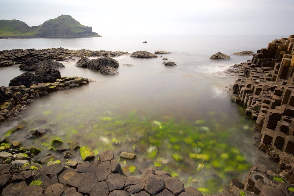 A rocky coastal landscape with a calm sea, green moss-covered rocks, and a distant island.