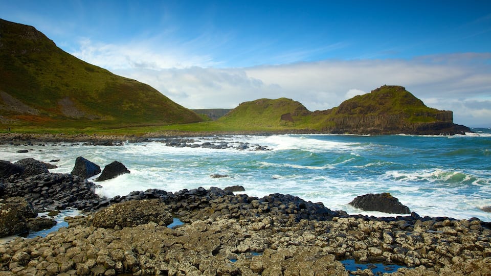 A rocky coastline with waves crashing against the shore, green hills in the background, and a clear sky.