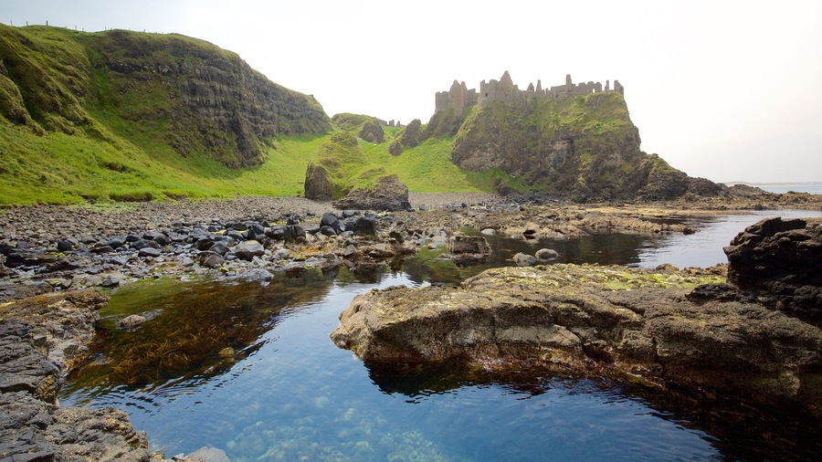 A rocky shoreline with clear water, green hills, and a castle ruin.