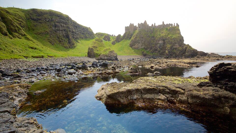 A rocky shoreline with clear water, green hills, and a castle ruin.