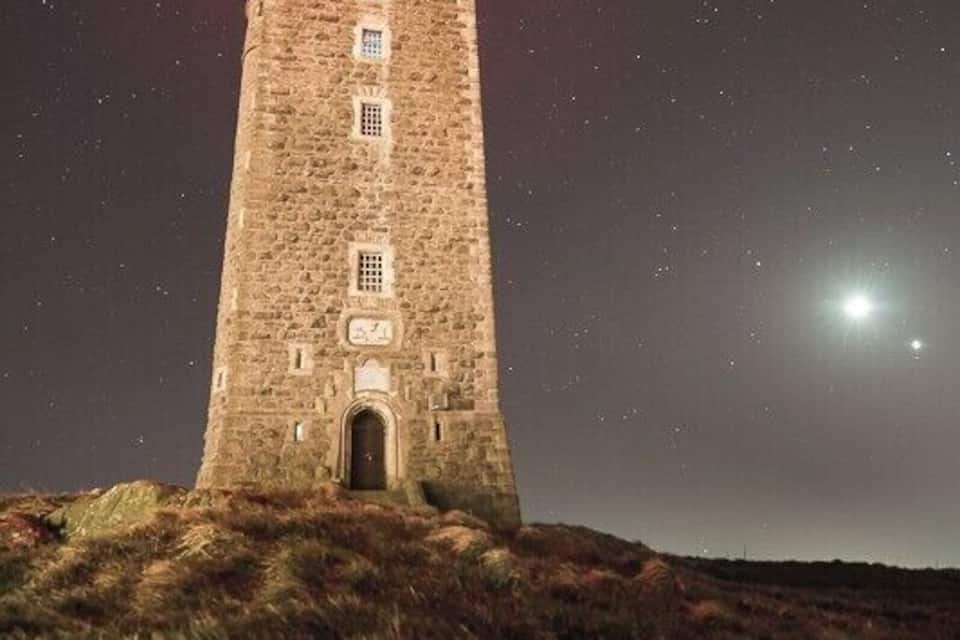 Uma torre de pedra com uma luz vermelha no topo, situada numa colina verdejante sob um céu estrelado.
