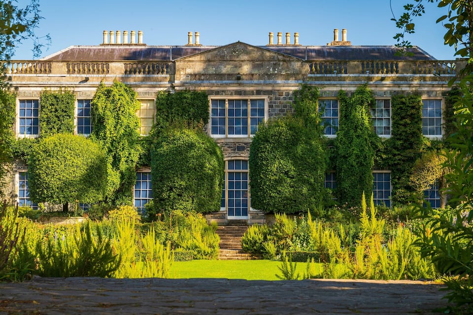 A large, historic building with a symmetrical facade, surrounded by lush greenery and a well-maintained lawn.