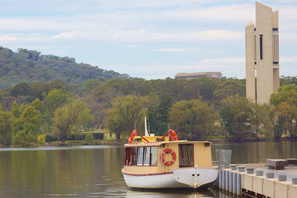 A river with a boat docked at a pier, surrounded by trees and a tall building in the background.