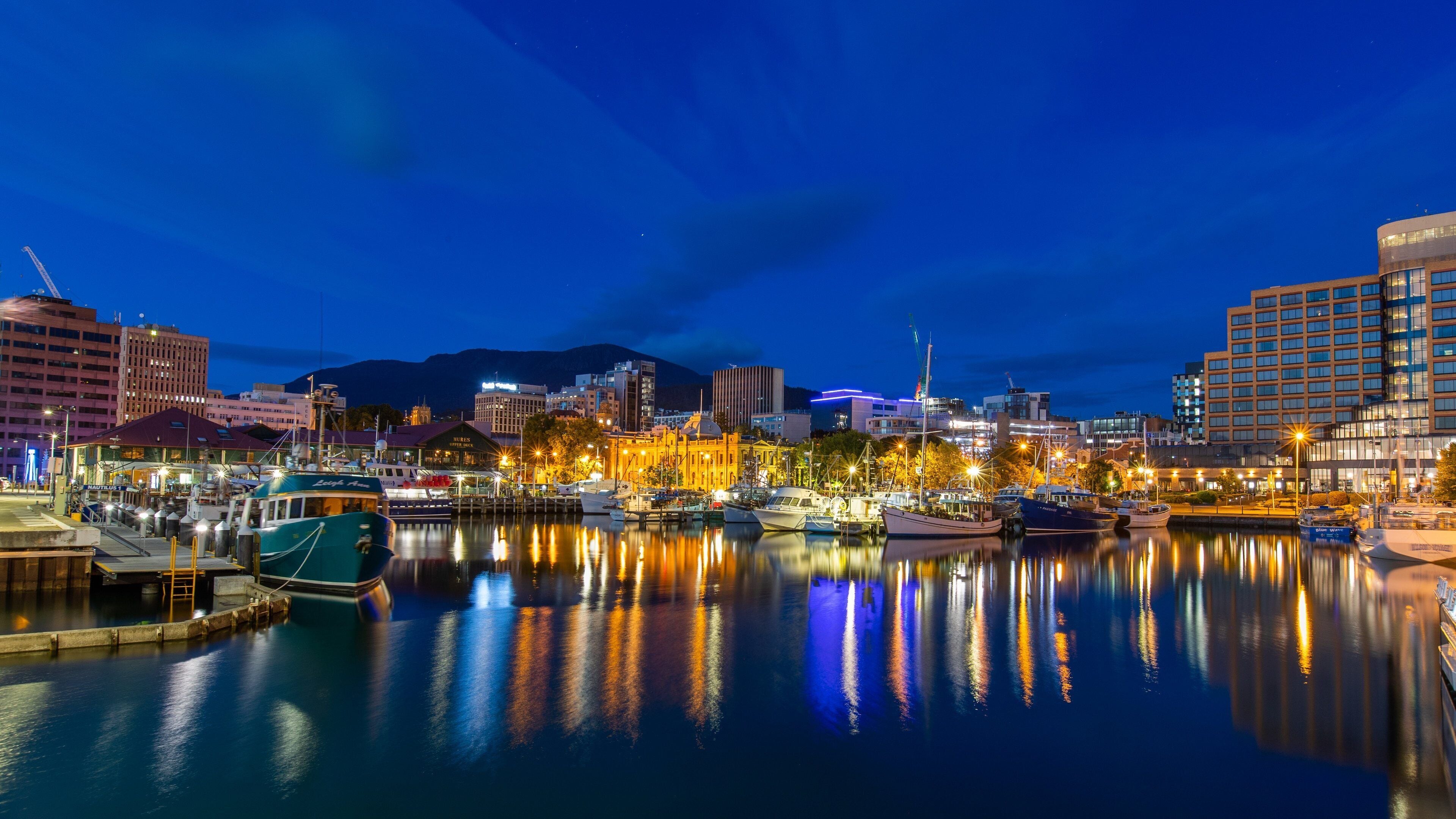 A marina at dusk with boats docked, reflecting city lights and buildings.