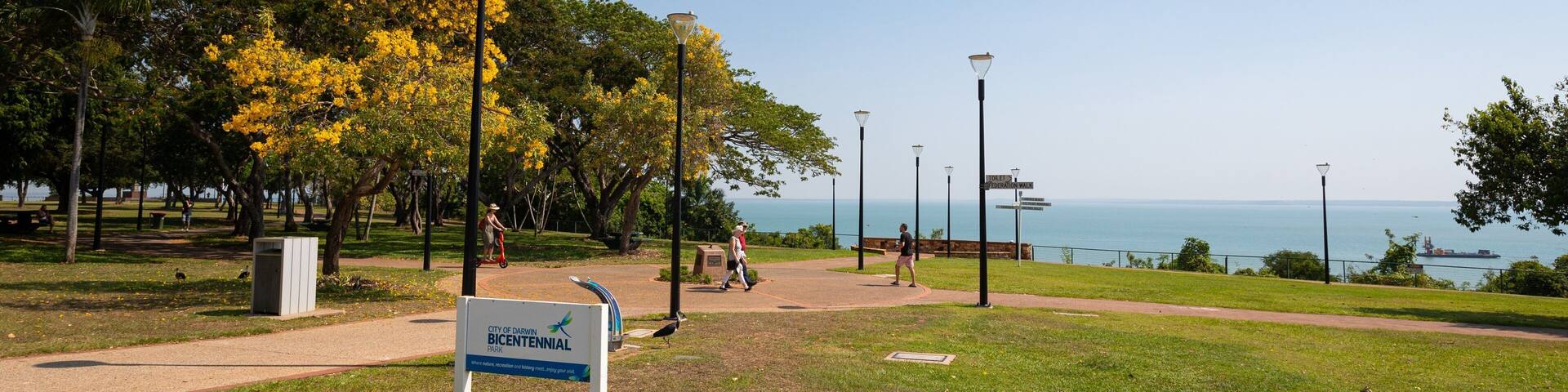 A park with a sign for the City of Darwin Bicentennial Park, a walking path, and a view of the ocean.
