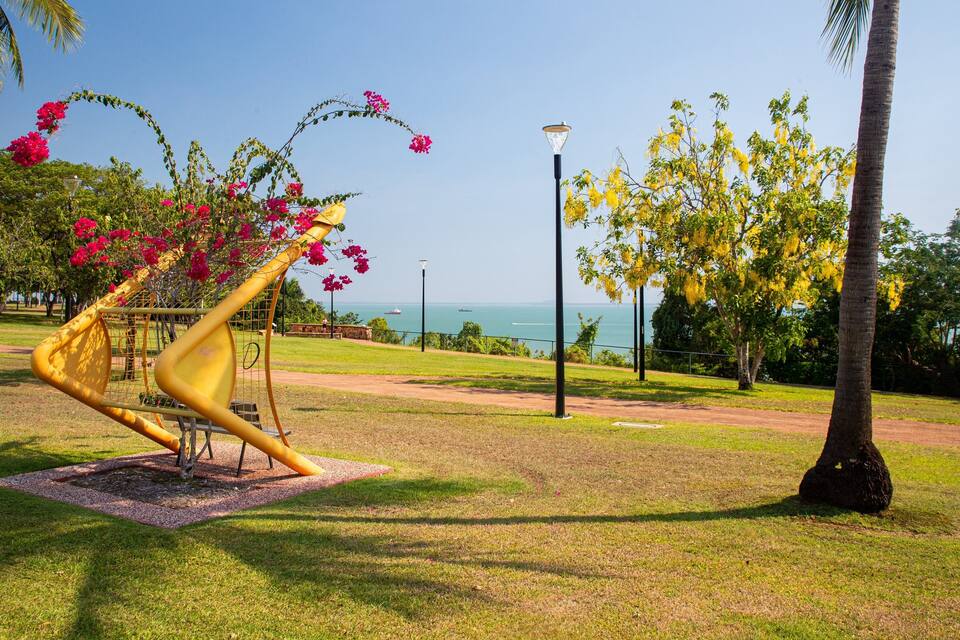 Een park met een gouden sculptuur, roze bloemen, gele bomen en uitzicht op de oceaan.