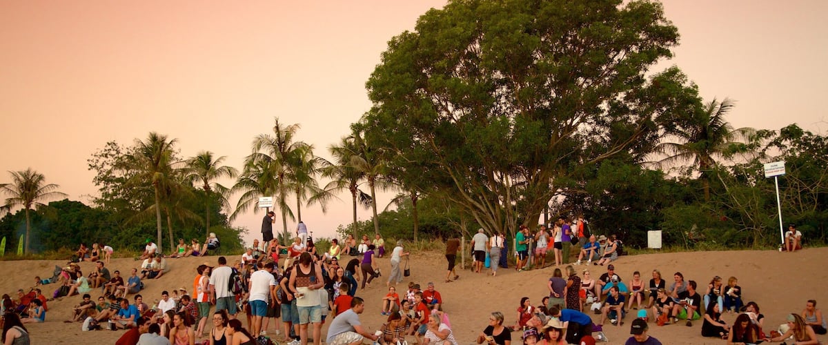 A crowded beach with palm trees and people sitting and standing on the sand.