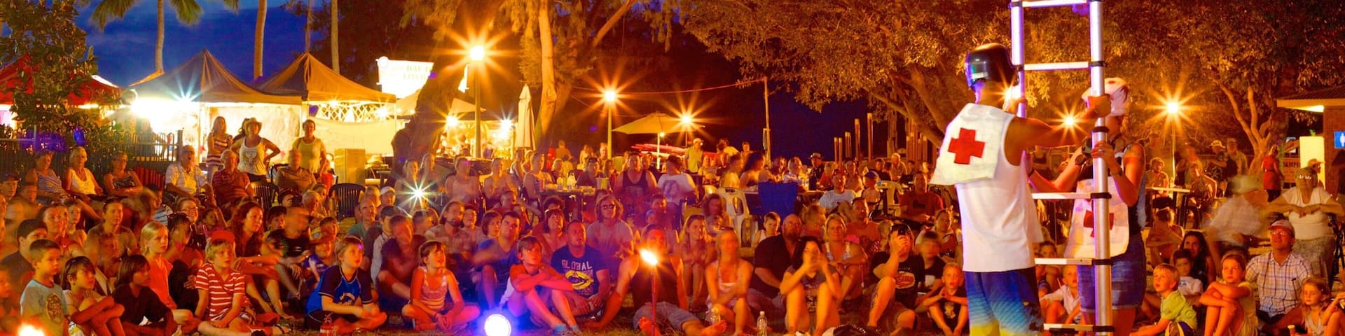 A fire performance with a performer on a tall ladder, surrounded by an audience in an outdoor setting with palm trees and tents.