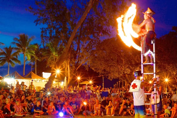Un spectacle de jonglage avec le feu, avec un artiste évoluant sur une grande échelle, entouré d’un public dans un cadre extérieur agrémenté de palmiers et de tentes.